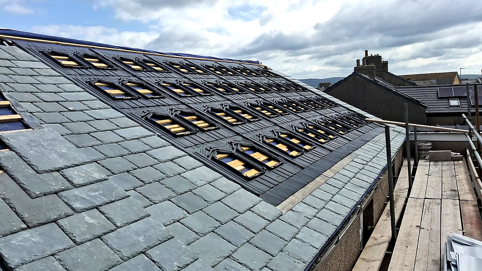 Wide view of the mounting trays set into the new slate across the whole roof plane, scaffold to one side and the village behind
