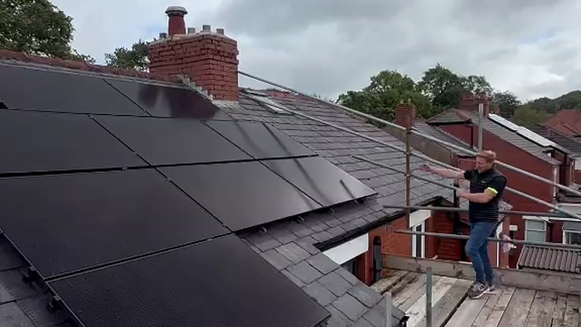 The finished array on the rear roof at Somerset Avenue, Wilpshire, filmed from the scaffold at handover: nine all black panels on a slate roof beside a brick chimney, one of the team on the scaffold pointing them out